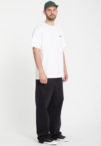 Hvit T-skjorte med en liten svart logo, kombinert med løstsittende svarte jeans og svarte kanvas sneakers, supplert med en grønn caps.