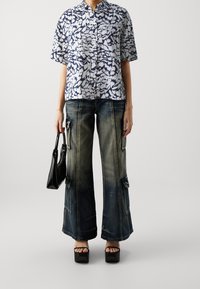Blusa a maniche corte, blu e bianca con motivo, dotata di colletto rialzato, abbinata a pantaloni in denim larghi e strappati con tasche e dettagli metallici.