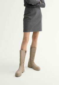 Bottes beiges jusqu'aux genoux avec une semelle plate et texturée et une fermeture éclair sur le côté, associées à une jupe ajustée à carreaux dans des tons de gris.