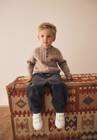 Child wearing a light brown knitted sweater with a cable design and buttons, paired with dark corduroy pants and white Velcro shoes, seated on a patterned trunk.