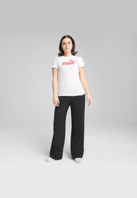 Maglietta bianca a maniche corte con logo rosso "PUMA", abbinata a pantaloni neri a gamba larga e calzature bianche, su uno sfondo neutro.