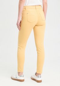 Jean skinny jaunes avec une texture lisse et deux poches arrière, associés à des baskets blanches avec des accents argentés et une semelle beige.
