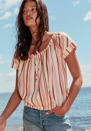 Nainen, jolla on märät tummat hiukset, pukeutunut raidalliseen lyhythihaiseen puseroon ja farkkushortseihin, seisoo meren rannalla puolipilvisen taivaan alla.