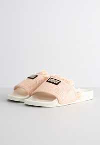 Pink woven slide sandals with fringe detail, a textured strap, and white rubber base. Features a black rectangular logo patch.