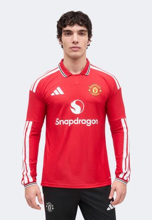 Mężczyzna w czerwonym długim rękawie koszulce piłkarskiej Manchester United z białymi paskami Adidas i logo Snapdragon, w połączeniu z czarnymi spodniami.