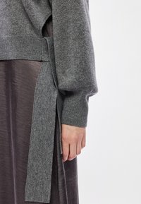 Γκρίζος πλεκτός σκούφος με λεπτομέρεια δέσμευσης στο πλάι, φέρει ραμμένο μανίκι. Μαλακή υφή και άνετη εφαρμογή, φορεμένος πάνω από μια μεταξένια σκούρα καφέ φούστα.