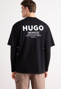 T-shirt in cotone nero con logo e testo "HUGO" bianchi sul retro, maniche corte e vestibilità rilassata. Tessuto uniforme, senza pattern.