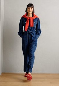 Macacão de ganga com mangas longas, cintura ajustada e bolsos de carga, combinado com um suéter vermelho atado ao pescoço e sandálias vermelhas.