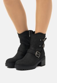 Botas negras de tobillo de ante con un tacón apilado, que cuentan con dos correas ajustables con hebilla y un zipper lateral para facilitar su uso.