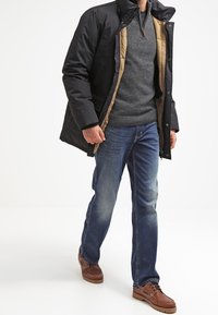 Manteau noir rembourré sur un pull gris, jeans bleus et chaussures marron. Le manteau a une doublure beige contrastante et un col zippé.