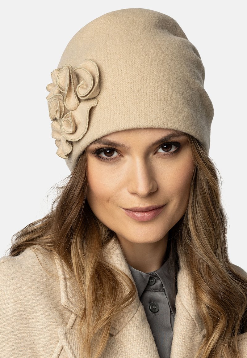 Bonnet en laine beige avec un détail de fleur en tissu superposé sur le côté. Texture lisse, forme arrondie, ajustement confortable.
