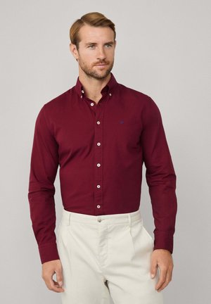 Chemise boutonnée bordeaux en coton avec des manches longues, boutons contrastés et un petit logo bleu sur la poitrine. Associée à un pantalon crème.