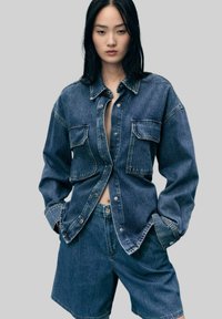 Chemise en denim, bleu foncé, avec deux poches poitrine, des boutons et une coupe décontractée ; associée à un short en denim assorti. Texture lisse.