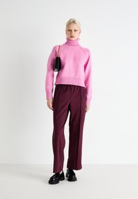 Pink knitted turtleneck sweater, ribbed hem, paired with maroon trousers featuring side zippers and black platform shoes. Black handbag with chain.