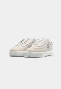 O pereche de sneakers Calvin Klein cu talpa joasă, bej și alb, cu șireturi albe și logo CK pe laterale, pe un fundal alb.