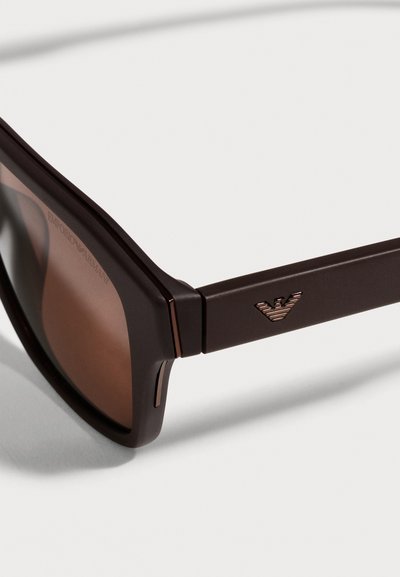 Gros plan sur des lunettes de soleil Emporio Armani brun foncé, avec des verres bruns et un logo d’aigle en métal sur la branche.