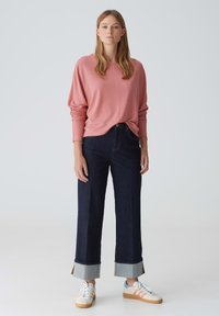 Rosa Langarmshirt, dunkelblaue weite Jeans mit umgeschlagenen Säumen und weiße Sneaker mit pastellfarbenen Streifen und einer braunen Sohle.