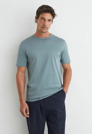 Man met een lichtblauw T-shirt met korte mouwen en donkere broek staat tegen een witte muur met één hand in zijn zak.