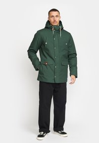 Groene waterdichte jas met een capuchon, drukknoop- en ritssluiting, voortassen en contrasterende bruin leren accenten. Zwarte broek en schoenen.