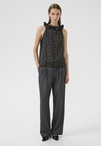 Schwarze ärmellose Bluse mit Rüschensaum und weißen Punkten, kombiniert mit grauen weiten Hosen und spitzen schwarzen Schuhen.