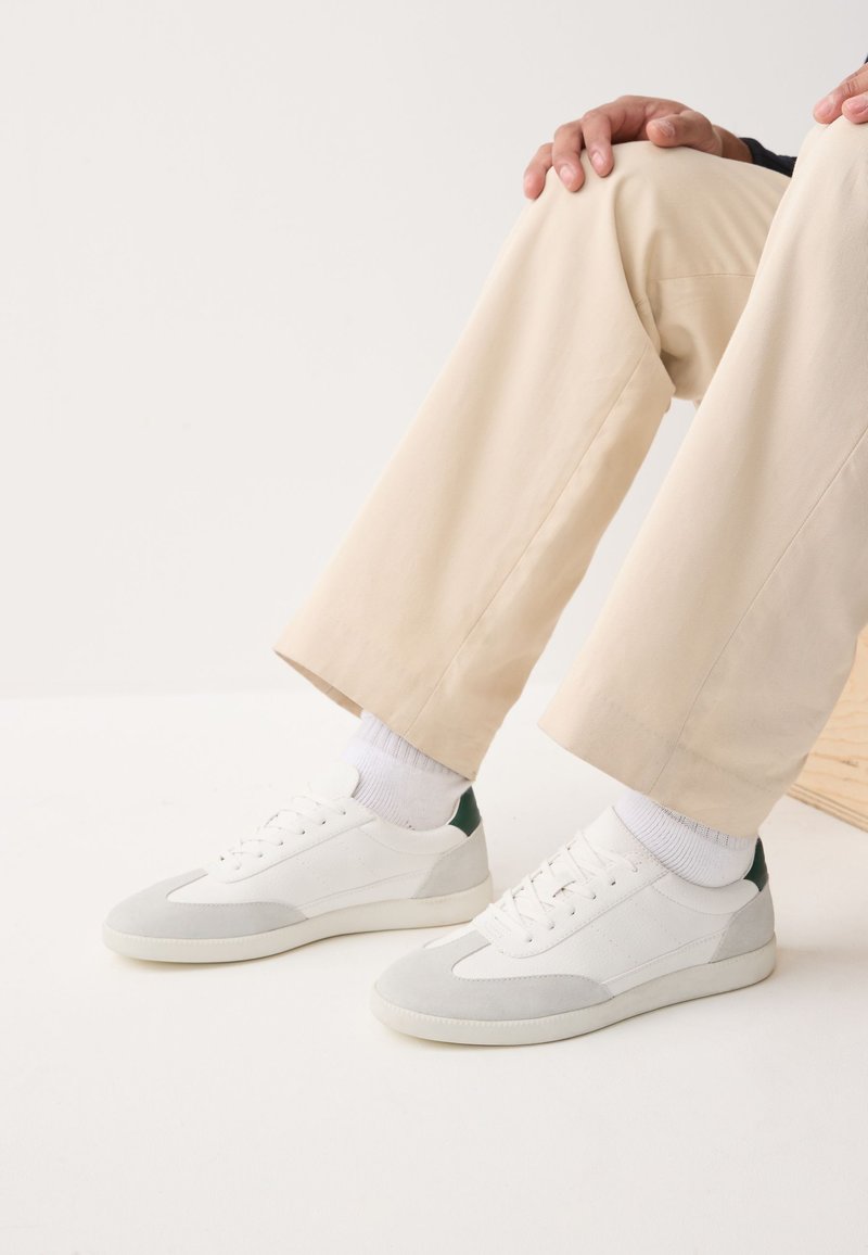 Personne assise, les mains reposant sur le genou, portant un pantalon beige, des chaussettes blanches et des baskets blanches avec des accents gris et vert foncé.