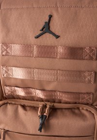 Barna textúrájú hátizsák három vízszintes varrott pánttal, fekete Jumpman logóval, és dupla cipzárral a front zsebén.