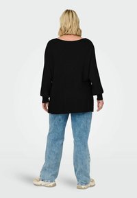 En person med axellångt blont hår vänder ryggen mot kameran och bär en lös svart tröja och ljusblå jeans i en minimalistisk miljö.
