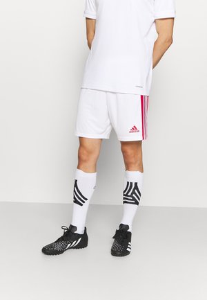 Mann, der weiße Adidas-Sportshorts mit roten Streifen, weiße Socken mit schwarzen Mustern und schwarze Fußballschuhe trägt, steht vor weißem Hintergrund.