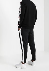 Sweat-shirt noir avec des rayures rouges et blanches à motifs sur les manches, associé à un pantalon de jogging noir avec des bandes blanches sur les côtés, porté avec des baskets blanches.