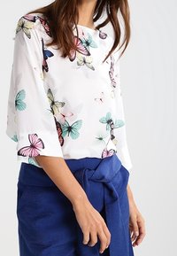 Blouse blanche avec un imprimé coloré de papillons, dotée de manches larges et d'un col rond, associée à une jupe portefeuille bleue taille haute.