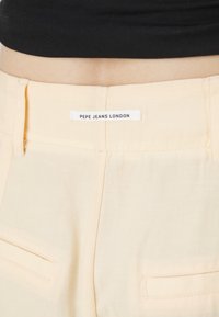 Světlé žluté kalhoty s hladkým povrchem, obsahující bílou nášivku s nápisem "PEPE JEANS LONDON" a dvěma zadními kapsami.