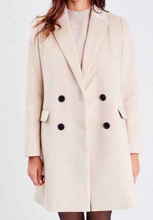 Femme portant un manteau en laine beige croisé avec des boutons noirs, par-dessus un pull en tricot beige clair, debout devant un fond uni.