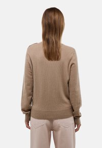 Personne aux cheveux bruns lisses portant un pull beige et un pantalon beige clair, vue de dos sur un fond clair uni.