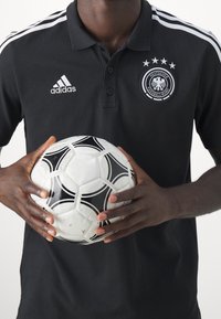Černé polo tričko Adidas s bílými pruhy a logem, na kterém je emblém Německé fotbalové asociace. Ruce drží černobílý fotbal.