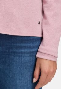 Helles pinkfarbiges, texturiertes Strickpullover mit hochgestellten Ärmeln, kombiniert mit dunkelblauen Jeans. Verfügt über einen kleinen Markenakzent nahe dem Saum.