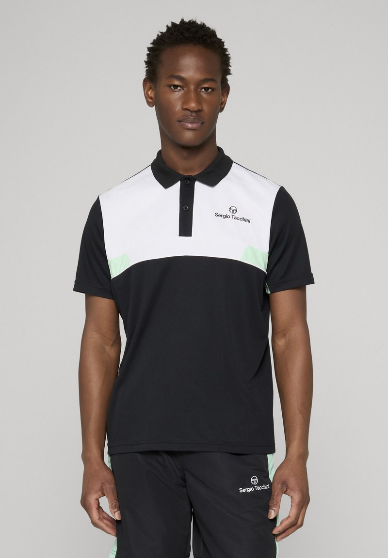 Man met een zwart-wit Sergio Tacchini poloshirt en korte broek, staand tegen een effen lichtgrijze achtergrond.