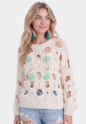Vrouw met lang blond haar draagt een crème gebreide trui met grote gaten en groene bloemdetails, gecombineerd met gescheurde denimshorts en oorbellen met veren.