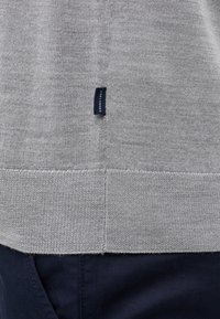 Maglione grigio lavorato a maglia con una finitura texturizzata, caratterizzato da una piccola etichetta blu sul lato e un orlo dritto. Abbinato a pantaloni blu navy.