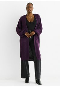 Long, open-front cardigan in deep purple knit fabric with oversized sleeves and two front pockets, worn over a black top and dark pants.