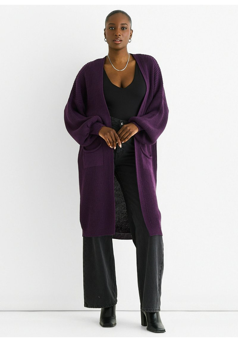 Long, open-front cardigan in deep purple knit fabric with oversized sleeves and two front pockets, worn over a black top and dark pants.
