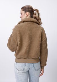 Nő copfba kötött hajjal, barna sherpa dzsekit és világoskék farmert viselve, oldalra néz, hátának egy része a kamera felé fordul.