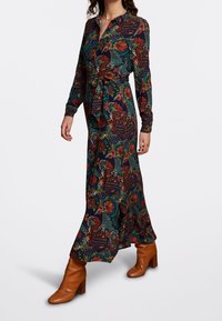 Kvinna som bär en långärmad maxi-klänning med blommönster och midjeband, tillsammans med bruna läderstövlar med klack som går till vaden.