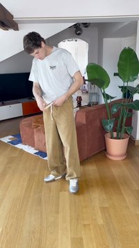 T-shirt grigio chiaro e pantaloni in velluto a coste beige con coulisse. In piedi su un pavimento di legno, sneakers argentate, foglie verdi sullo sfondo.