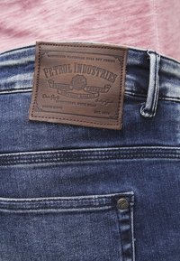Mörkblå denimjeans med en texturerad brun läderlapp märkt "Petrol Industries." Funktioner inkluderar sömnad och en silverfärgad knappdetalj.
