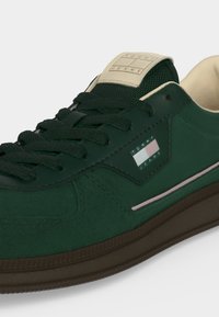 Σκούρο πράσινο αθλητικό παπούτσι Tommy Jeans με μαύρα κορδόνια, καφέ σόλα, μπεζ ετικέτα στη γλώσσα και λογότυπο Tommy Jeans στο πλάι.