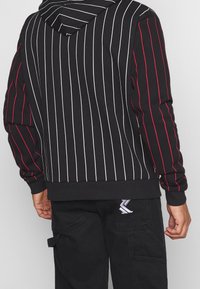 Musta huppari, jossa on pystysuorat valkoiset ja punaiset raidat, huppu ja resorireuna. Selässä on kontrastinen merkki mustissa housuissa.