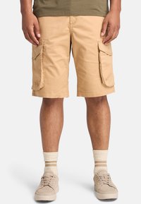 Pantaloni cargo beige in cotone, dotati di due grandi tasche laterali, con una vestibilità rilassata e orli rifiniti. Indossati con sneaker beige e calzini a righe.