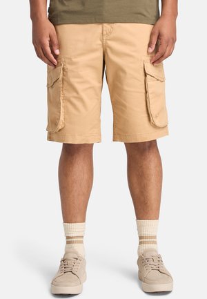 Pantalones cortos cargo beiges de algodón, con dos grandes bolsillos laterales, un corte relajado y bordes dobladillos. Llevados con zapatillas beiges y calcetines a rayas.