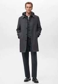 Abrigo de lana gris con frente abotonado, gran cuello y bolsillos laterales. Combinado con una camiseta oscura y jeans negros, usado con zapatos negros.