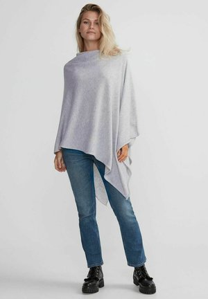 Ljusgrå poncho i mjukt material, med en lös, asymmetrisk design. Matchad med blå jeans och svarta snörstövlar.